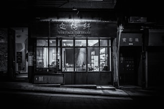 A dimly lit, monochrome exterior of a tea house with a sign that reads 'Heritage Tea House' above the entrance. The large front windows display part of the interior, showing tables and chairs. The establishment appears closed, with minimal light and no visible patrons.