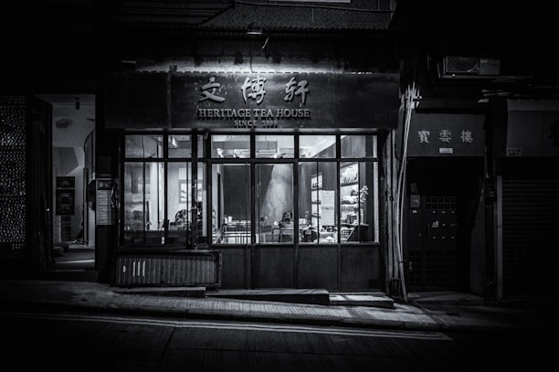 A dimly lit, monochrome exterior of a tea house with a sign that reads 'Heritage Tea House' above the entrance. The large front windows display part of the interior, showing tables and chairs. The establishment appears closed, with minimal light and no visible patrons.