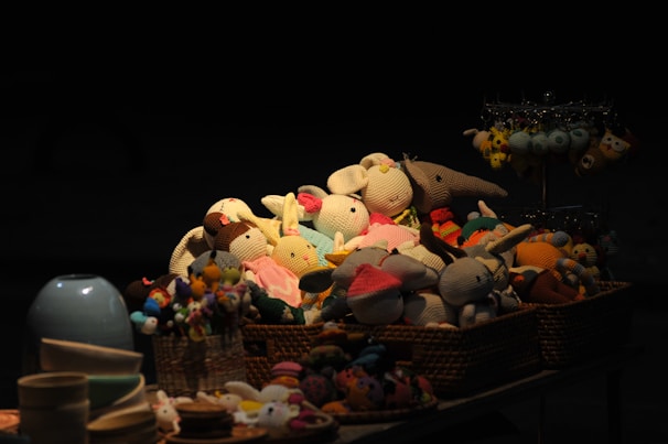 A collection of hand-knitted stuffed animals is arranged in wicker baskets, brightly colored and illuminated against a dark background. The toys include various animals wearing cute outfits, giving a cozy and inviting feel. Nearby, there are other plush toys on a display stand, as well as some ceramic bowls in the foreground.