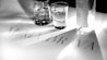Black and white image featuring a glass of water and a bottle on a tabletop. The surface has the text 'ALMA BRAVA' with light creating shadows.