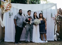 A group of four people pose together in front of a decorative backdrop featuring flowers and greenery. Two of the individuals appear to be a bride and groom, with the bride holding a bouquet of white flowers. The groom holds a small black and white dog. The backdrop is adorned with white curtains and floral arrangements.