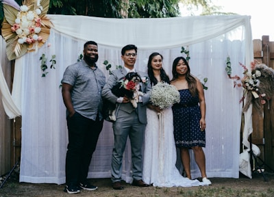 A group of four people pose together in front of a decorative backdrop featuring flowers and greenery. Two of the individuals appear to be a bride and groom, with the bride holding a bouquet of white flowers. The groom holds a small black and white dog. The backdrop is adorned with white curtains and floral arrangements.