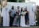 A group of four people pose together in front of a decorative backdrop featuring flowers and greenery. Two of the individuals appear to be a bride and groom, with the bride holding a bouquet of white flowers. The groom holds a small black and white dog. The backdrop is adorned with white curtains and floral arrangements.