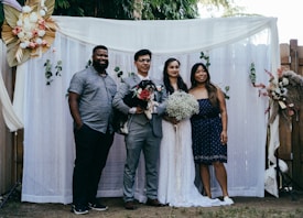 A group of four people pose together in front of a decorative backdrop featuring flowers and greenery. Two of the individuals appear to be a bride and groom, with the bride holding a bouquet of white flowers. The groom holds a small black and white dog. The backdrop is adorned with white curtains and floral arrangements.