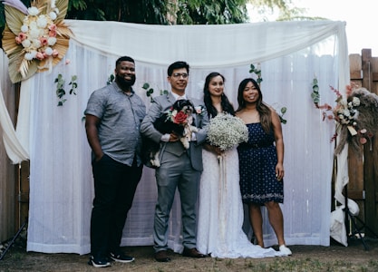 A group of four people pose together in front of a decorative backdrop featuring flowers and greenery. Two of the individuals appear to be a bride and groom, with the bride holding a bouquet of white flowers. The groom holds a small black and white dog. The backdrop is adorned with white curtains and floral arrangements.