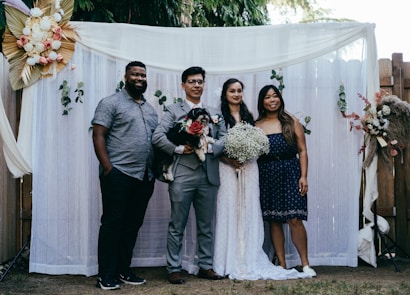 A group of four people pose together in front of a decorative backdrop featuring flowers and greenery. Two of the individuals appear to be a bride and groom, with the bride holding a bouquet of white flowers. The groom holds a small black and white dog. The backdrop is adorned with white curtains and floral arrangements.