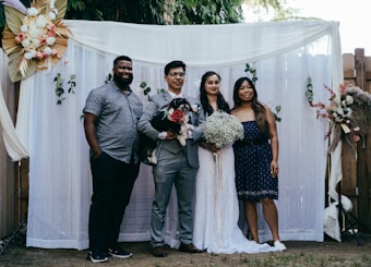 A group of four people pose together in front of a decorative backdrop featuring flowers and greenery. Two of the individuals appear to be a bride and groom, with the bride holding a bouquet of white flowers. The groom holds a small black and white dog. The backdrop is adorned with white curtains and floral arrangements.