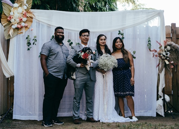 A group of four people pose together in front of a decorative backdrop featuring flowers and greenery. Two of the individuals appear to be a bride and groom, with the bride holding a bouquet of white flowers. The groom holds a small black and white dog. The backdrop is adorned with white curtains and floral arrangements.