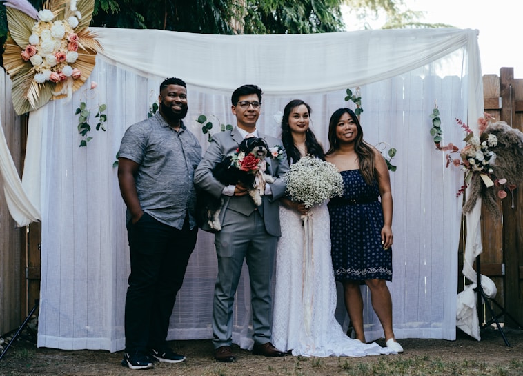 A group of four people pose together in front of a decorative backdrop featuring flowers and greenery. Two of the individuals appear to be a bride and groom, with the bride holding a bouquet of white flowers. The groom holds a small black and white dog. The backdrop is adorned with white curtains and floral arrangements.