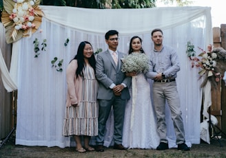 Four people are standing in front of a white draped backdrop with decorative flowers and greenery. Two individuals are in casual attire, while the other two appear to be a bride and groom, with the bride holding a bouquet of white flowers. The setting seems to be an outdoor event, possibly a wedding, given the attire and floral arrangements.