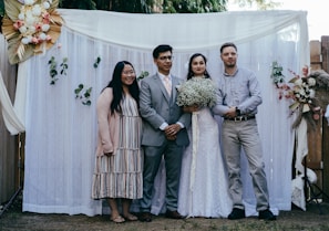 Four people are standing in front of a white draped backdrop with decorative flowers and greenery. Two individuals are in casual attire, while the other two appear to be a bride and groom, with the bride holding a bouquet of white flowers. The setting seems to be an outdoor event, possibly a wedding, given the attire and floral arrangements.