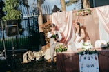 A couple is celebrating at an outdoor wedding reception. The man is wearing a vest and shirt and is cheerfully opening a bottle, causing the liquid to spray. The woman, dressed in a white wedding gown, smiles at the scene. The backdrop includes pink and rose gold drapery with floral arrangements. A cake and other decorations such as photographs are on the table, and a speaker is visible in the background.