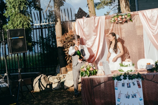 A couple is celebrating at an outdoor wedding reception. The man is wearing a vest and shirt and is cheerfully opening a bottle, causing the liquid to spray. The woman, dressed in a white wedding gown, smiles at the scene. The backdrop includes pink and rose gold drapery with floral arrangements. A cake and other decorations such as photographs are on the table, and a speaker is visible in the background.