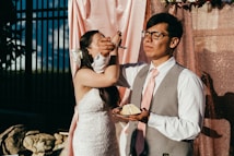 A couple is standing in front of a pink and shimmering backdrop. The man is wearing a suit with a pink tie and glasses, holding a plate with a piece of cake. The woman, dressed in a white wedding gown, is playfully feeding him a bite using a fork. Sunlight casts shadows against the backdrop, creating a warm atmosphere.
