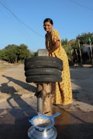 A woman in a yellow patterned dress is operating a manual water pump in an outdoor rural setting. She is smiling and looking at the camera while the pump dispenses water into a metal pot through a blue sieve. Two recycled tires are repurposed above the pump, possibly for structural support or as part of the design. The background includes a few trees and a simple structure made of logs.