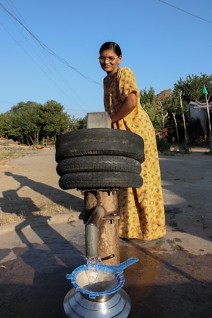 A woman in a yellow patterned dress is operating a manual water pump in an outdoor rural setting. She is smiling and looking at the camera while the pump dispenses water into a metal pot through a blue sieve. Two recycled tires are repurposed above the pump, possibly for structural support or as part of the design. The background includes a few trees and a simple structure made of logs.
