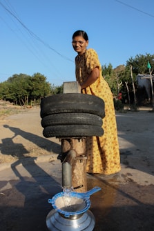 A woman in a yellow patterned dress is operating a manual water pump in an outdoor rural setting. She is smiling and looking at the camera while the pump dispenses water into a metal pot through a blue sieve. Two recycled tires are repurposed above the pump, possibly for structural support or as part of the design. The background includes a few trees and a simple structure made of logs.