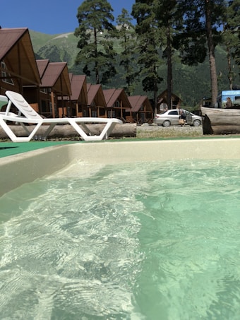 A series of wooden cabins with brown roofs are lined up in the background, surrounded by tall pine trees. In the foreground, clear water possibly from a hot tub or pool is visible with a white lounge chair nearby. A car is parked on the gravel area beside the cabins, with picturesque green mountains under a clear blue sky as the backdrop.