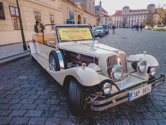 A vintage-style convertible car is parked on a cobblestone street in a historical area. The car is decorated with a sign offering sightseeing tours for 50 euros or 1200 CZK. There are buildings with classic architecture in the background along with a few pedestrians and parked cars.