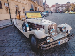 A vintage-style convertible car is parked on a cobblestone street in a historical area. The car is decorated with a sign offering sightseeing tours for 50 euros or 1200 CZK. There are buildings with classic architecture in the background along with a few pedestrians and parked cars.