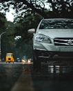 A silver car is parked on a wet road, with trees in the background casting shadows overhead. In the distance, a yellow auto rickshaw and other vehicles are visible on the road, with headlights creating small flares. The scene is set in an urban environment with a moody, overcast sky.