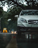 A silver car is parked on a wet road, with trees in the background casting shadows overhead. In the distance, a yellow auto rickshaw and other vehicles are visible on the road, with headlights creating small flares. The scene is set in an urban environment with a moody, overcast sky.