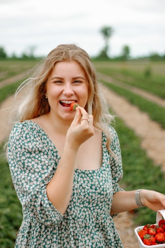 A woman with long blonde hair and a green floral dress is smiling while holding a strawberry near her mouth. She stands in a strawberry field with rows of plants behind her, and she is holding a container filled with strawberries.