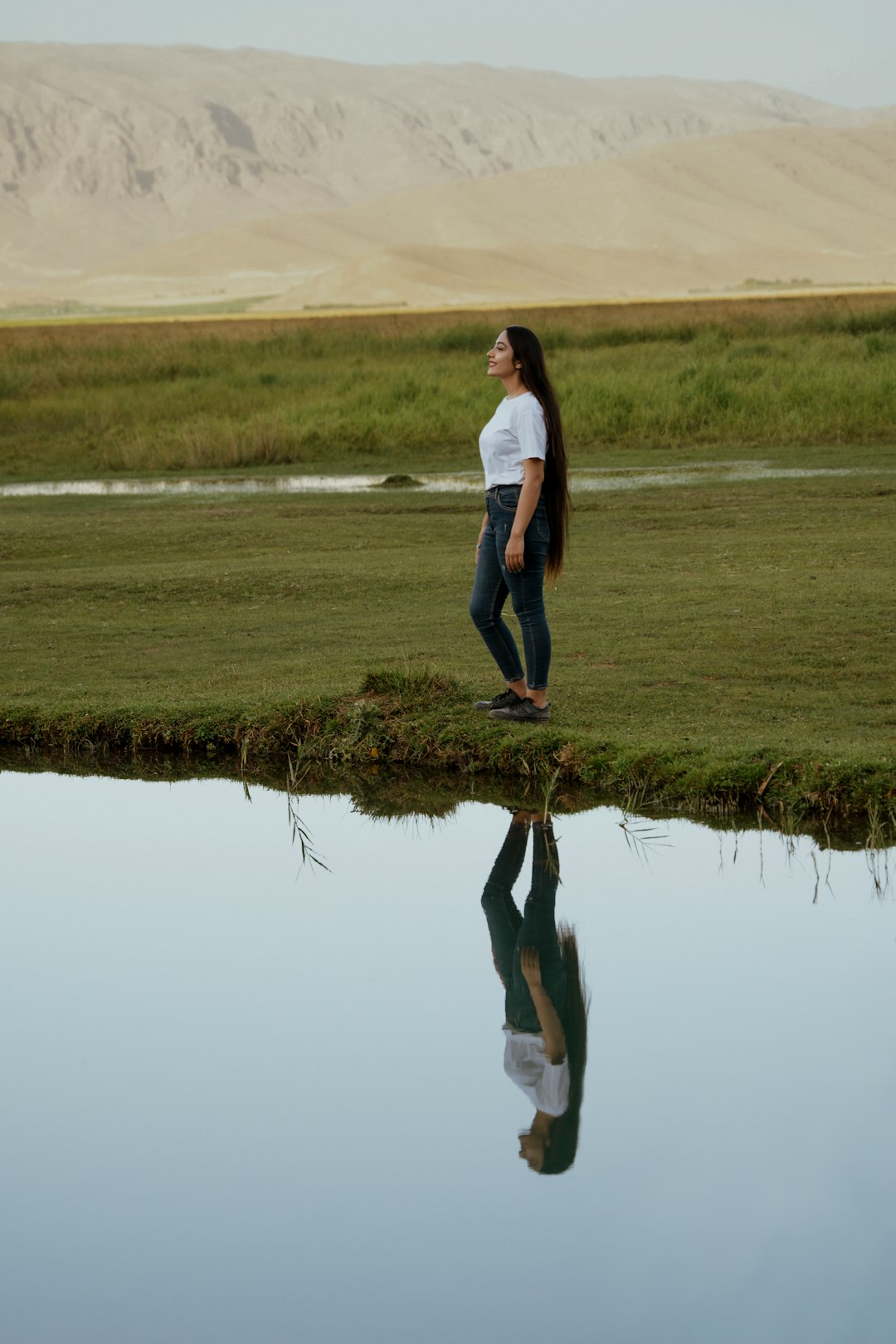woman in white shirt and black pants standing on green grass field near lake during daytime