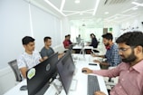 A group of people working at computers in an office, sitting in a row. The space appears modern with white walls, sleek furniture, and bright lighting. Some individuals have headphones on, indicating a focused or collaborative environment.