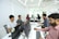 A group of people working at computers in an office, sitting in a row. The space appears modern with white walls, sleek furniture, and bright lighting. Some individuals have headphones on, indicating a focused or collaborative environment.