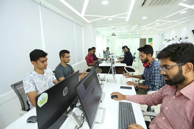 A group of people working at computers in an office, sitting in a row. The space appears modern with white walls, sleek furniture, and bright lighting. Some individuals have headphones on, indicating a focused or collaborative environment.