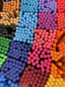 A collection of colorful marker pens arranged neatly in a display. The pens are stacked closely together in groups based on color, forming a visually appealing pattern. Various colors such as blue, red, orange, yellow, green, pink, and purple are represented.