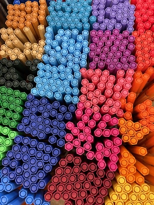 A collection of colorful marker pens arranged neatly in a display. The pens are stacked closely together in groups based on color, forming a visually appealing pattern. Various colors such as blue, red, orange, yellow, green, pink, and purple are represented.