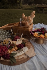 A picnic spread is arranged on a blanket featuring a wicker basket filled with baguettes and a bottle of rose. A platter displays an assortment of cheeses, crackers, grapes, and olives, garnished with herbs. Next to it, a tray holds vibrant fruit slices, including melons and berries. In the background, lush greenery and a wooden bench can be seen.