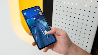 A person is holding a smartphone displaying a mobile game with vibrant graphics. The game depicts a character running on train tracks. The background includes a well-lit wall with a pattern of holes.