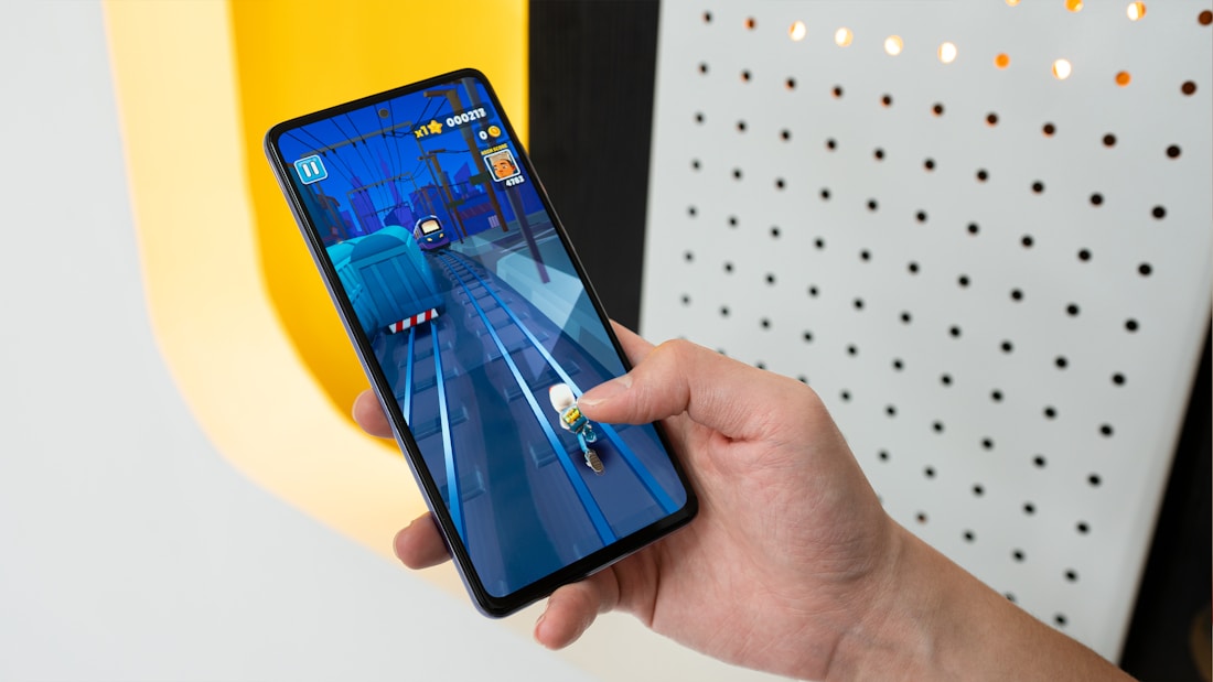 A person is holding a smartphone displaying a mobile game with vibrant graphics. The game depicts a character running on train tracks. The background includes a well-lit wall with a pattern of holes.