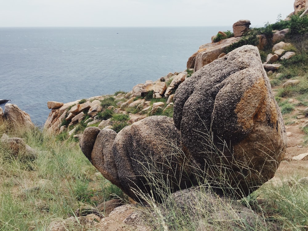 uomo in giacca nera che si siede sulla formazione rocciosa marrone vicino allo specchio d'acqua durante il giorno