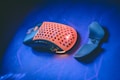 A gaming mouse with a perforated orange top surface and black base sits on a vibrant blue surface. Next to it lies a detachable thumb rest attachment.