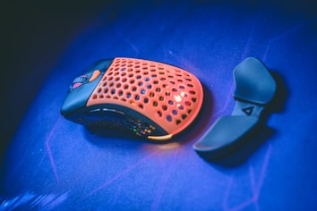A gaming mouse with a perforated orange top surface and black base sits on a vibrant blue surface. Next to it lies a detachable thumb rest attachment.