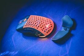A gaming mouse with a perforated orange top surface and black base sits on a vibrant blue surface. Next to it lies a detachable thumb rest attachment.