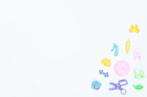 Various colorful plastic toys are scattered on a pale background. The toys include a yellow crab, a pink parasol, a green rocking horse, a purple pair of scissors, and a light blue elephant among others.