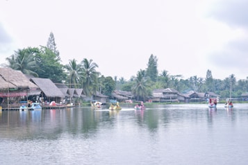 A serene lakeside scene featuring traditional thatched-roof huts along the water's edge. Paddle boats shaped like animals are being used by people enjoying leisure activities on the calm lake. Tall palm trees and lush greenery frame the background, creating a tranquil and inviting atmosphere.