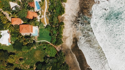 An aerial view of a coastal area featuring lush greenery and several houses with orange-tiled roofs. Next to the houses, a bright blue swimming pool is visible. The scene is bordered by a sandy beach leading to the ocean, where waves are crashing onto the shore.