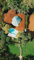 Aerial view of a residential property featuring a house with a red-tiled roof and a distinctive blue and white polygonal roof accent, surrounded by lush greenery. A small, kidney-shaped swimming pool with a nearby lounge area featuring white sunbeds and an umbrella is visible. The garden has a well-manicured lawn, various trees, and a stone pathway.