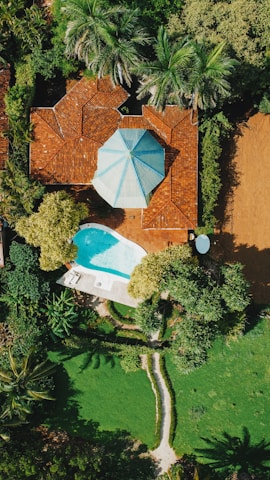Aerial view of a residential property featuring a house with a red-tiled roof and a distinctive blue and white polygonal roof accent, surrounded by lush greenery. A small, kidney-shaped swimming pool with a nearby lounge area featuring white sunbeds and an umbrella is visible. The garden has a well-manicured lawn, various trees, and a stone pathway.