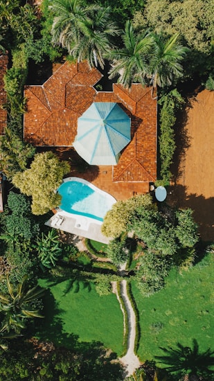 Aerial view of a residential property featuring a house with a red-tiled roof and a distinctive blue and white polygonal roof accent, surrounded by lush greenery. A small, kidney-shaped swimming pool with a nearby lounge area featuring white sunbeds and an umbrella is visible. The garden has a well-manicured lawn, various trees, and a stone pathway.