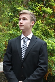 A young person dressed in a formal black suit with a striped tie, standing amidst lush green foliage. The background features vibrant green leaves and some orange berries, suggesting a garden or natural setting.