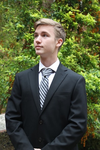 A young person dressed in a formal black suit with a striped tie, standing amidst lush green foliage. The background features vibrant green leaves and some orange berries, suggesting a garden or natural setting.