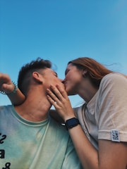 A couple is kissing under a clear blue sky, conveying a sense of intimacy and affection. The woman has long hair and wears a watch, while both display relaxed and comfortable poses.