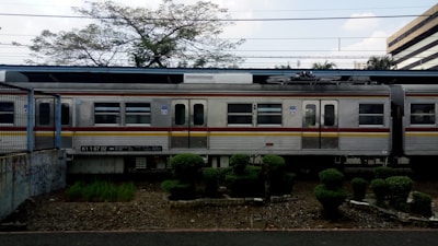A train with multiple carriages is seen stationary on a railway track. The train is predominantly silver with a red and yellow line running horizontally along its side. There is a patch of well-maintained garden with neatly trimmed bushes in front of the train. The background includes some trees with sparse leaves and a building to the far right.